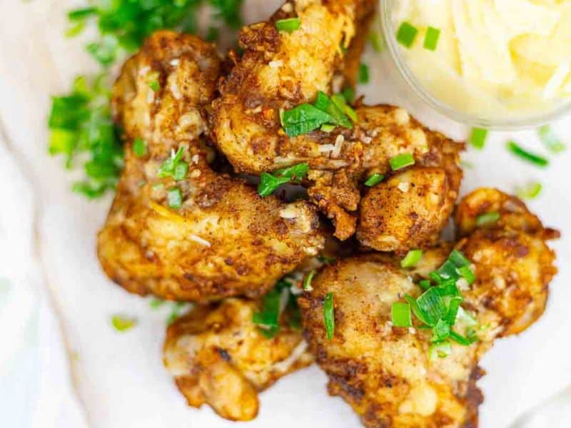 A plate of seasoned and baked chicken drumsticks garnished with chopped green herbs, served with a small dish of creamy sauce on the side.