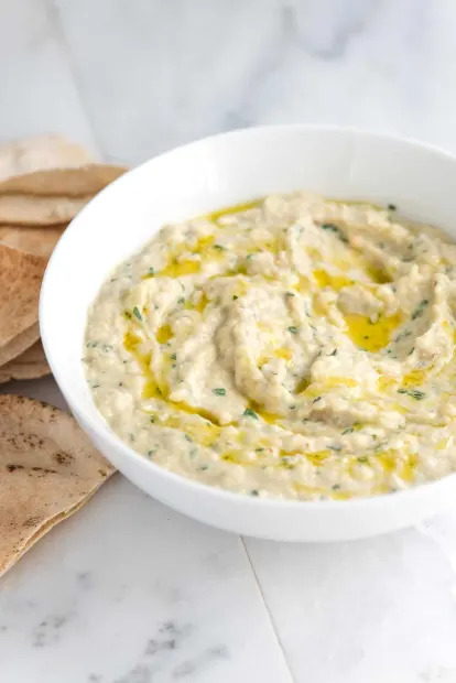 Bowl of baba ghanoush drizzled with olive oil, garnished with herbs, with pita bread on the side