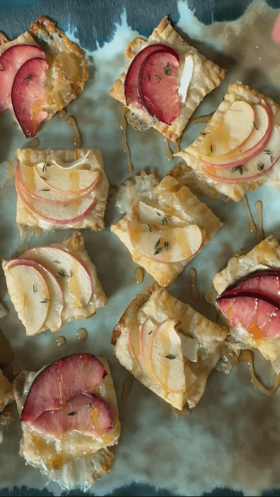 Square slices of pastry topped with apple, plum slices, thyme, and drizzled with honey on a teal surface.
