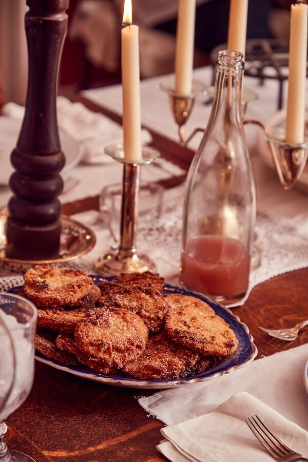 A plate of sugar-coated apple fritters sits on a table set with candles, a glass bottle of pink liquid, and neatly arranged white napkins, evoking sweet nostalgia and cozy memories.