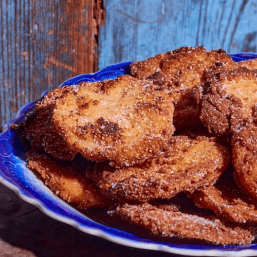 A blue plate filled with golden-brown, crispy apple fritters coated in sugar, set against a rustic wooden background, evokes warm nostalgia.