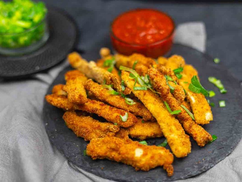 A plate of breaded and fried chicken strips garnished with herbs, served with a bowl of red dipping sauce in the background.