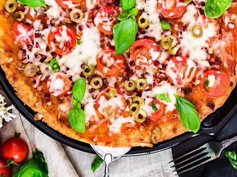 A baked pizza topped with melted cheese, tomato slices, green olives, and fresh basil leaves on a round black tray.