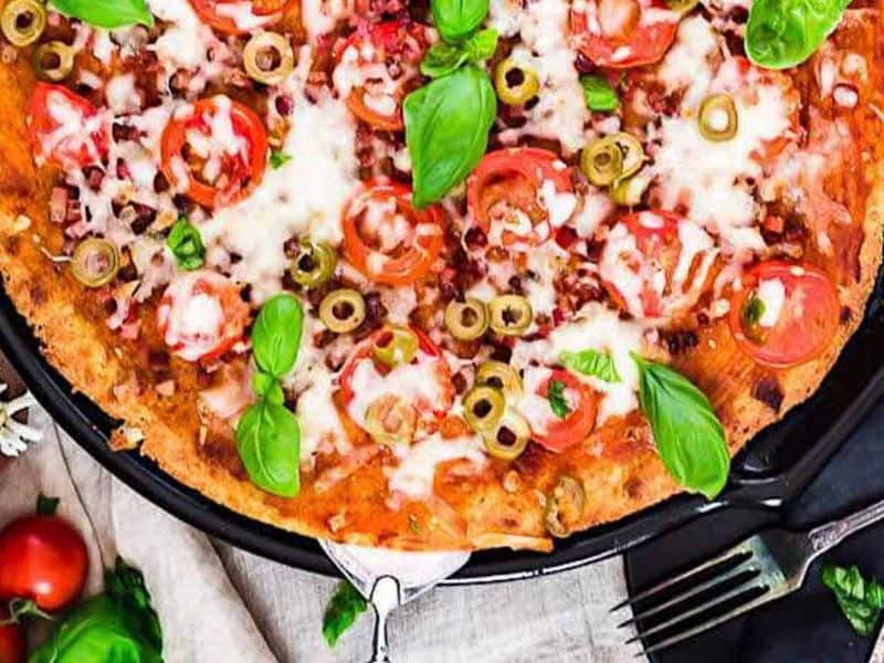 Close-up of a baked pizza topped with sliced tomatoes, green olives, melted cheese, fresh basil leaves, and visible herbs on a black pan. Fork and napkin placed beside the pizza.