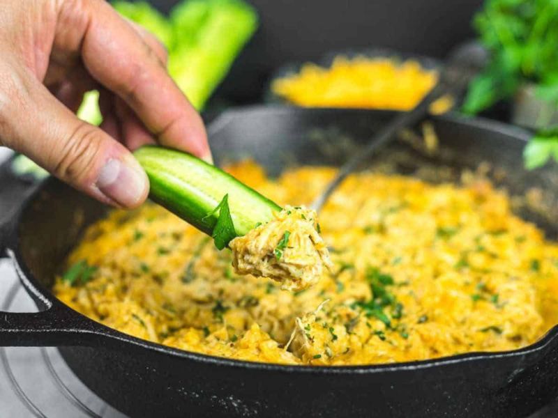 A hand dips a cucumber slice into a skillet filled with creamy, orange buffalo chicken dip garnished with chopped herbs.