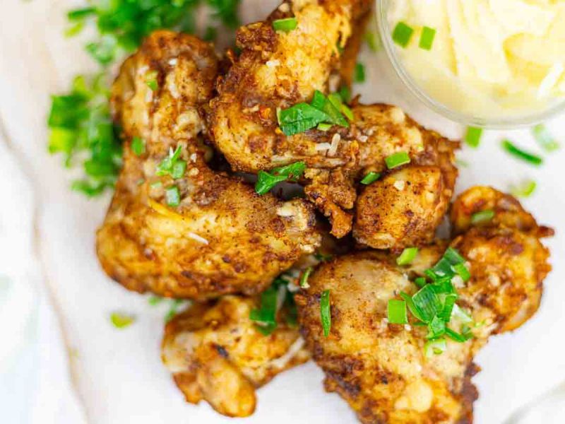 A plate of seasoned and baked chicken drumsticks garnished with chopped green herbs, served with a small dish of creamy sauce on the side.