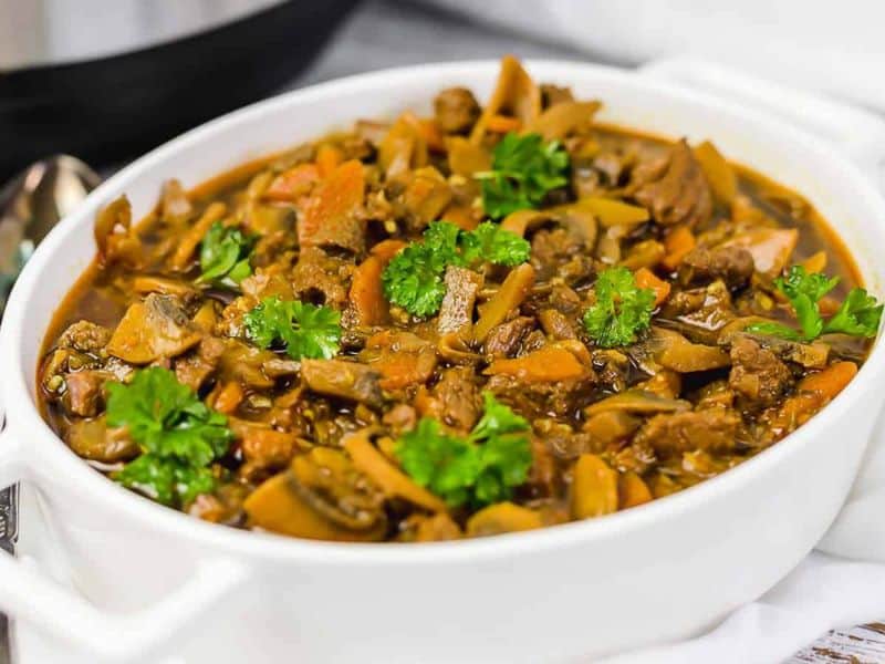 A white bowl filled with beef stew containing chunks of meat, carrots, mushrooms, and garnished with fresh parsley.