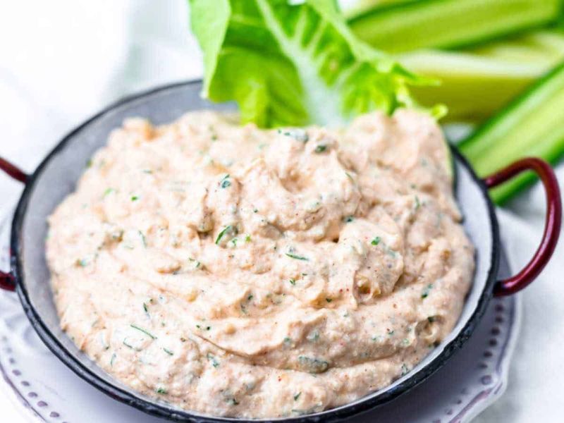 A bowl of creamy dip with visible herbs, served with fresh green celery sticks and lettuce in the background.