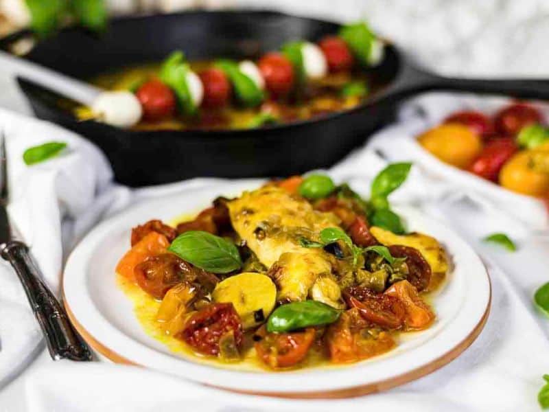 A plate of cooked chicken with tomatoes, yellow squash, and basil, set in front of a skillet and skewered caprese salad.
