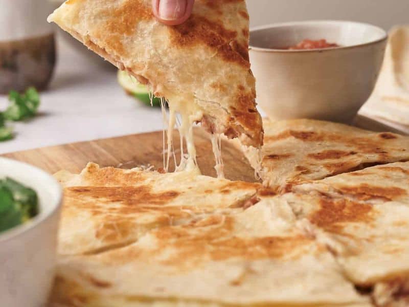 A hand lifts a cheesy quesadilla slice from a wooden board, with bowls of salsa and garnish in the background.