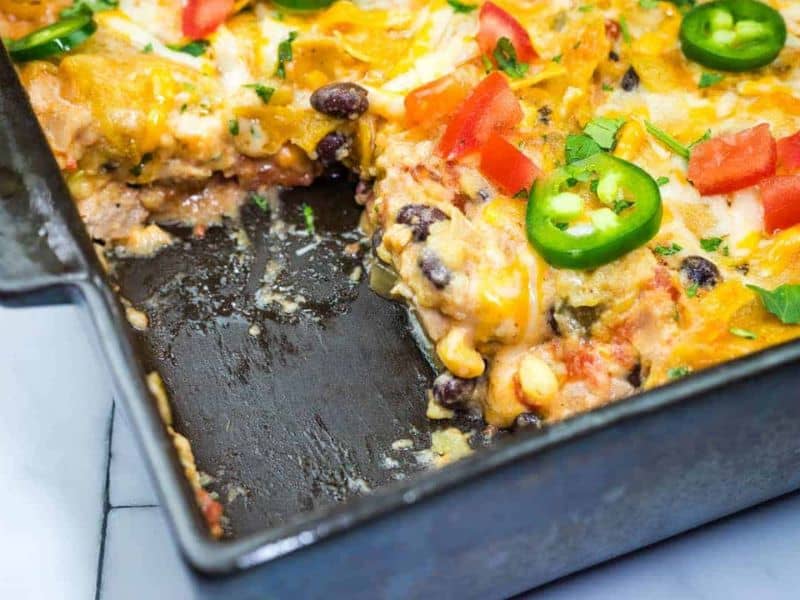 A casserole dish partially served, showing layers of beans, corn, tomatoes, cheese, and sliced jalapeños.