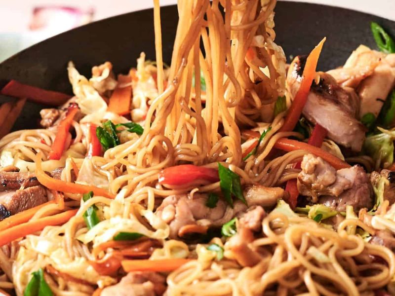 Close-up of stir-fried noodles with sliced chicken, carrots, bell peppers, cabbage, and green onions being lifted from a pan.