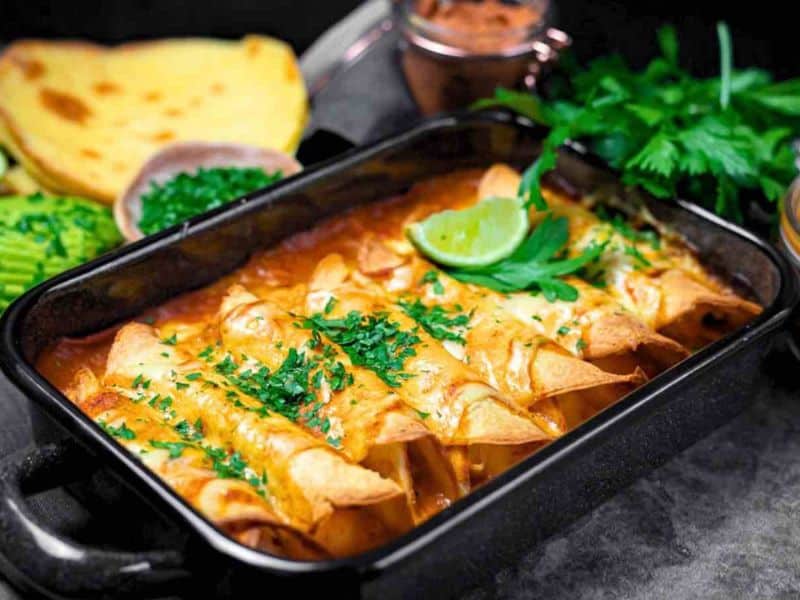 A baking dish with cheese-covered enchiladas garnished with chopped herbs and a lime wedge, with tortillas and green herbs in the background.