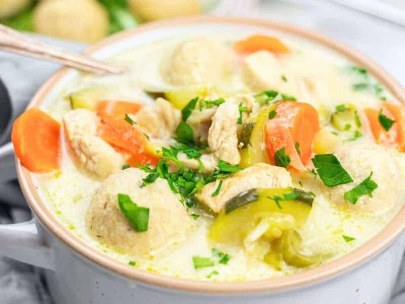 A bowl of creamy soup with chicken pieces, carrots, dumplings, and chopped herbs, garnished with fresh parsley.