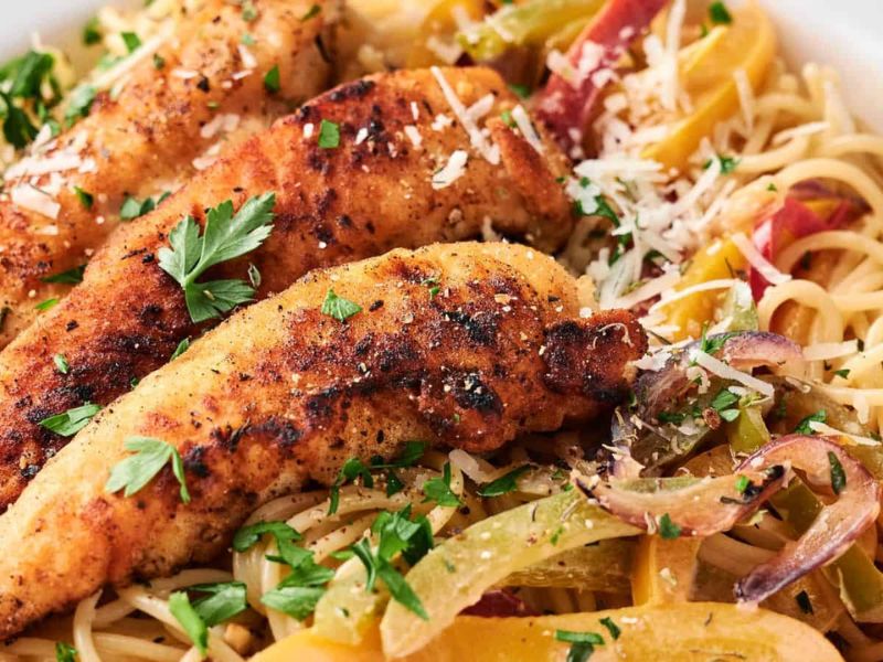 Close-up of seasoned, grilled chicken strips served over pasta with sliced bell peppers, onions, and garnished with chopped parsley and grated cheese.