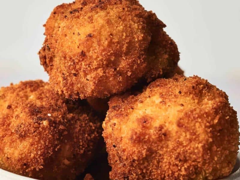 Three golden-brown, crispy breaded balls stacked on a white surface, likely fried and served as an appetizer or snack.