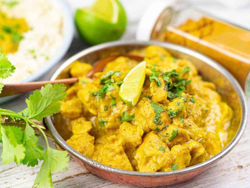 A bowl of chicken curry topped with cilantro and a lime wedge, with a jar of turmeric and a plate of rice in the background.