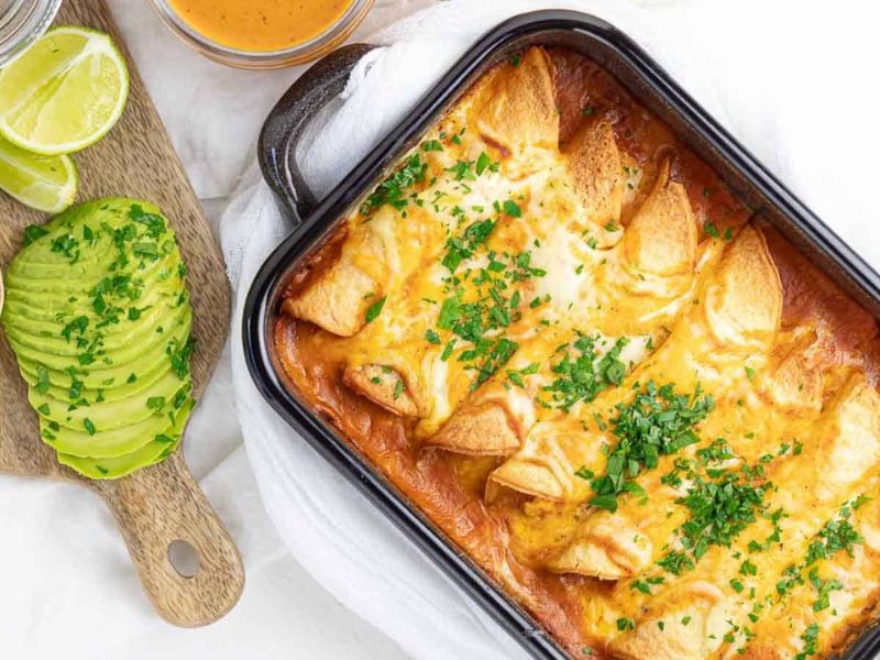 A tray of baked enchiladas topped with melted cheese and chopped herbs sits beside sliced avocado and lime wedges on a cutting board.