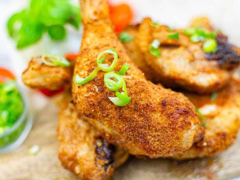 Close-up of crispy baked chicken drumsticks garnished with sliced green onions, served on a wooden surface with a side of green sauce and vegetables in the background.