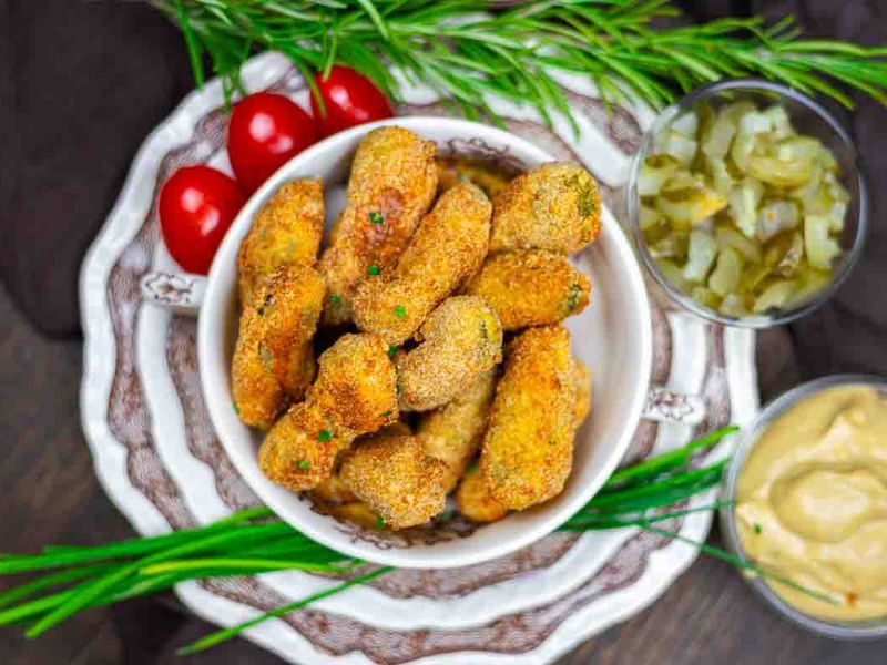 A bowl of breaded and fried pickle spears is surrounded by cherry tomatoes, chopped pickles, dipping sauce, and fresh herbs on stacked decorative plates.