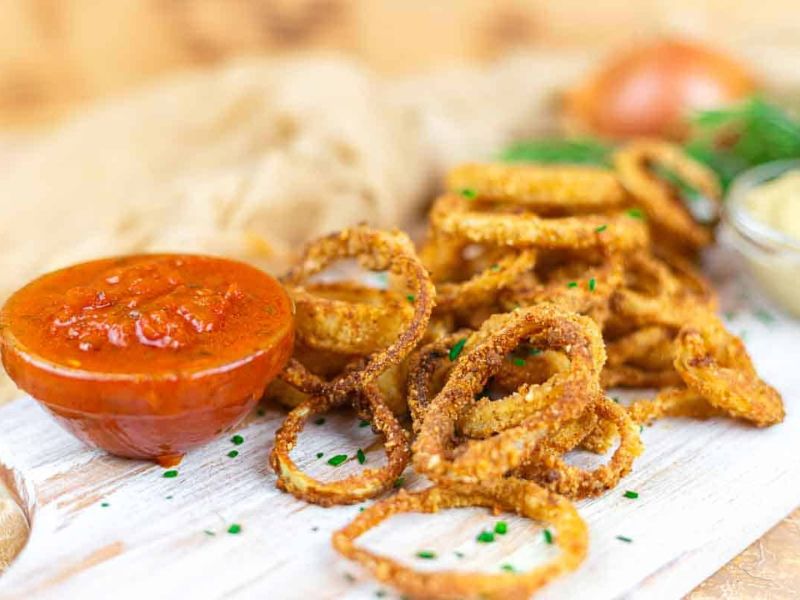 Crispy fried onion rings served on a white wooden board with a side of red dipping sauce and garnished with chopped chives.