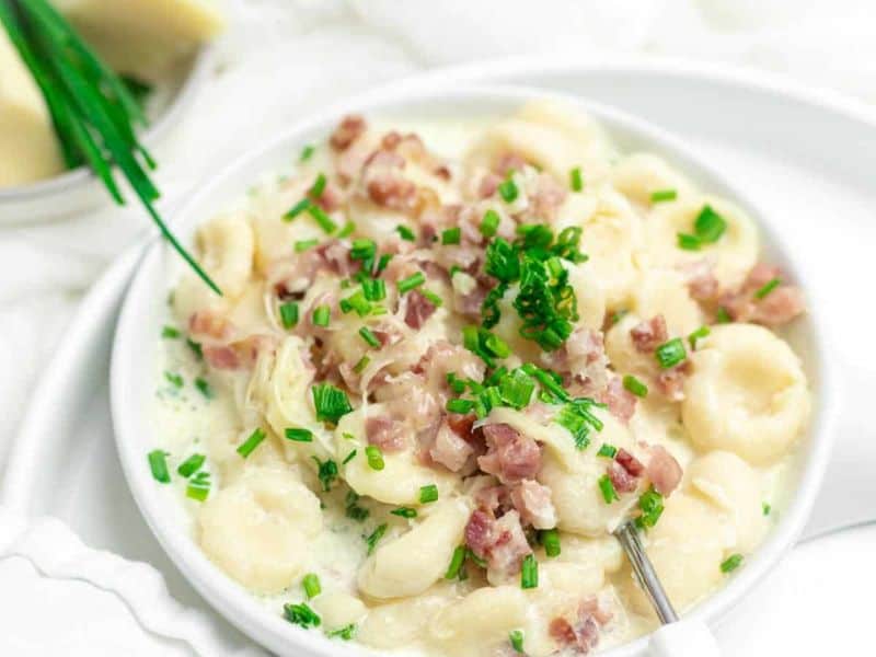 A bowl of creamy gnocchi topped with chopped bacon and fresh chives, served with a fork on a white plate.