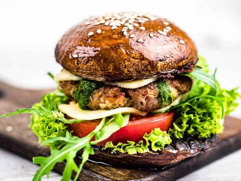 A burger with a portobello mushroom cap bun, lettuce, tomato, arugula, cheese, pesto, and a patty, served on a wooden board.