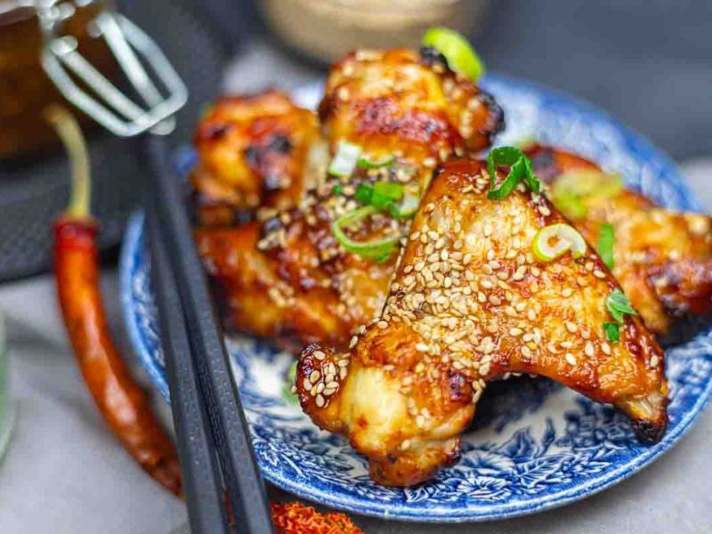 A plate of cooked chicken wings garnished with sesame seeds and chopped green onions, with chopsticks and a red chili pepper beside the plate.