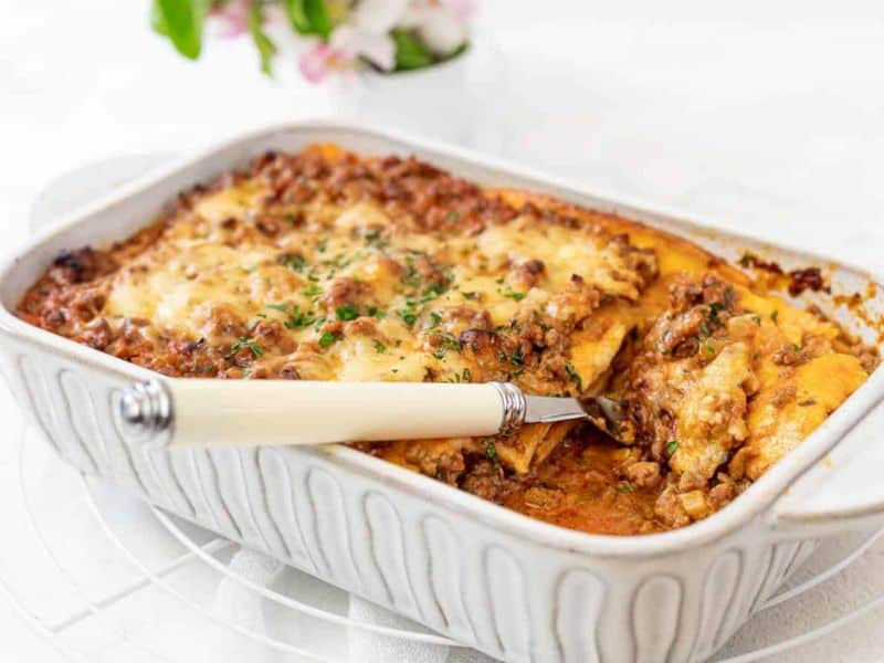 A baked dish of lasagna with a serving spatula lifting a portion, showing layers of pasta, meat sauce, and melted cheese in a white ceramic casserole dish.