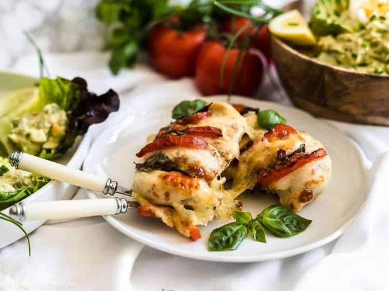 A plate with sliced baked chicken breast topped with melted cheese, tomato, and basil, served with a green salad.