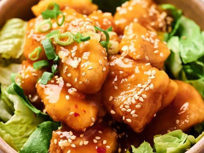 A bowl of crispy tofu cubes coated in sauce, topped with sesame seeds and sliced green onions, served on a bed of lettuce.