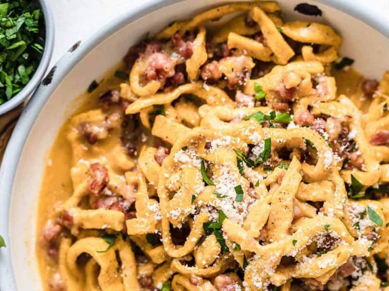 A close-up of a bowl of creamy pasta with pieces of bacon, grated cheese, and chopped parsley on top.