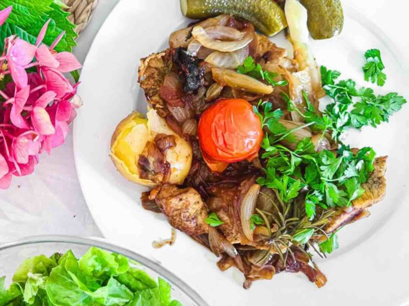 A white plate with roasted vegetables, including potatoes, onions, tomato, and herbs, garnished with parsley, next to pickles and a pink flower arrangement.