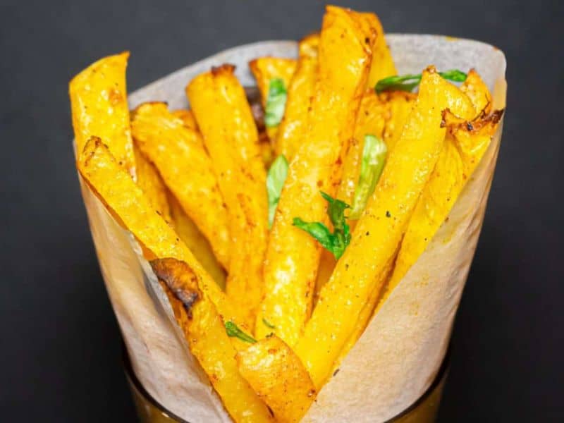 Golden baked fries served in a paper cone, garnished with fresh green herbs, set against a dark background.
