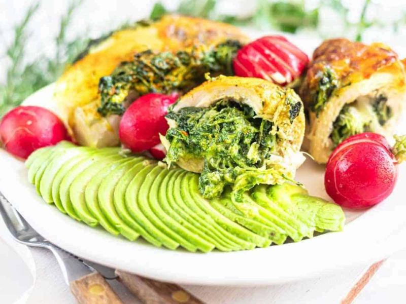 Sliced stuffed chicken breast with spinach, fresh radishes, and sliced avocado arranged on a white plate.