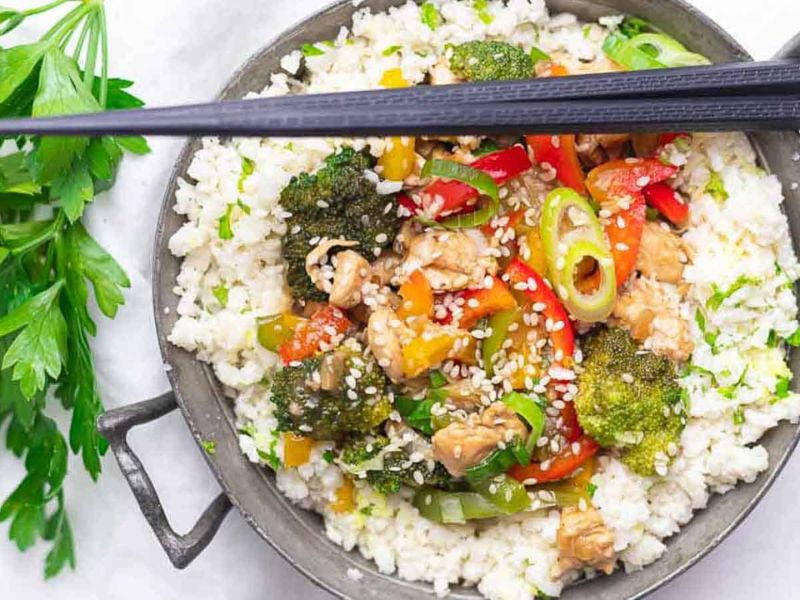 A bowl of stir-fried vegetables, chicken, and sesame seeds served over white rice, with chopsticks resting on top and parsley placed nearby.