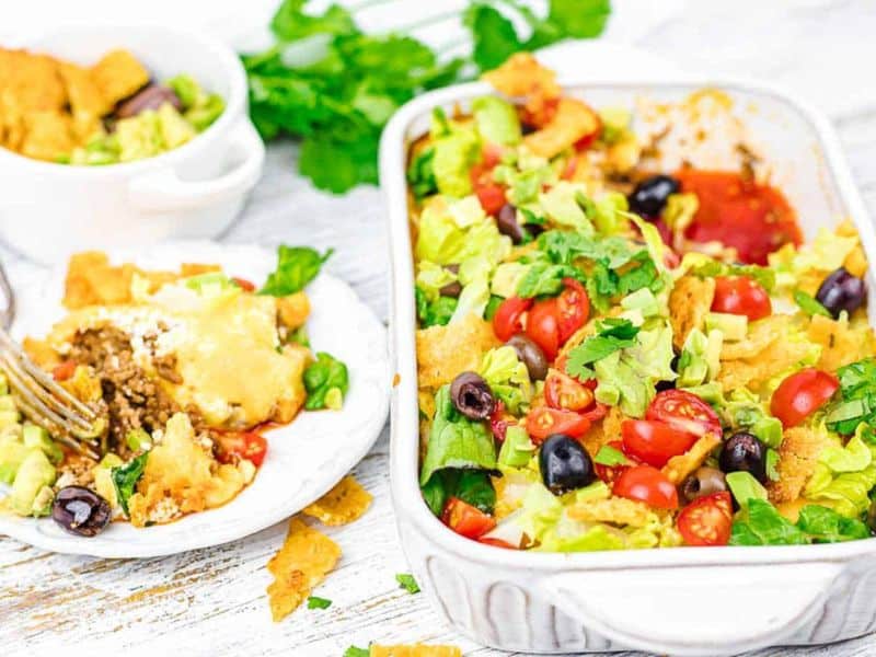 A casserole dish and plate with taco casserole topped with lettuce, tomatoes, olives, and cheese, alongside a bowl of guacamole on a white wooden surface.