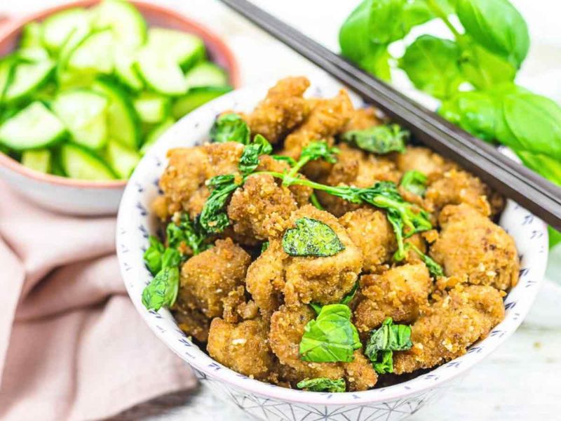 A bowl of crispy fried chicken pieces topped with fresh basil leaves, with a side dish of sliced cucumbers and a pair of chopsticks.