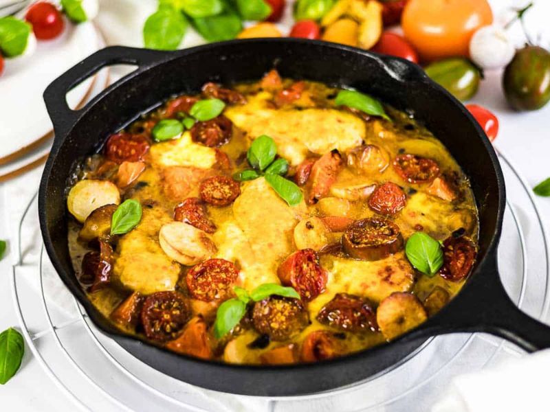 A cast iron skillet filled with baked chicken, tomatoes, garlic cloves, and basil leaves, surrounded by fresh vegetables on a white surface.