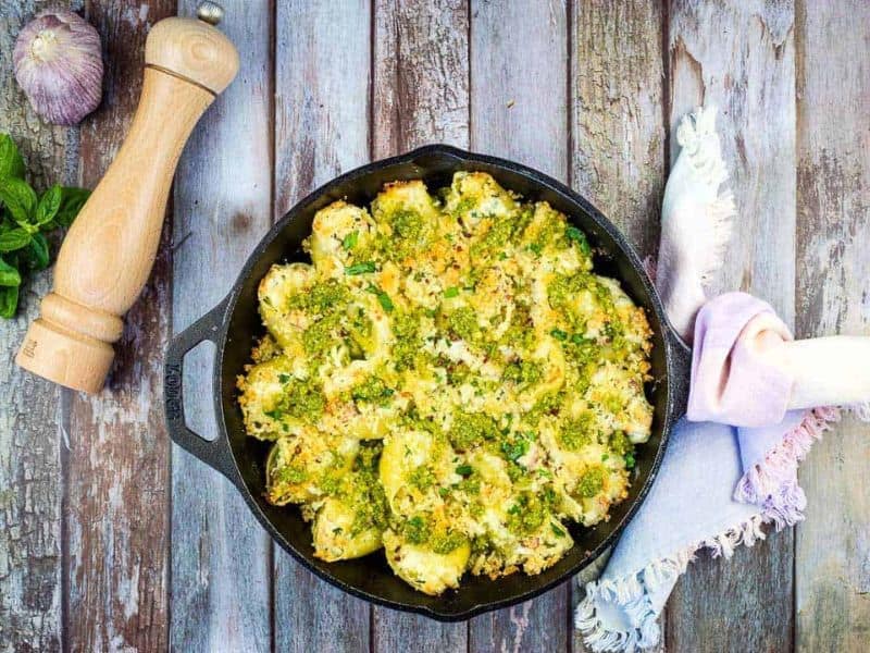 A cast iron skillet filled with baked stuffed pasta shells topped with herbs and breadcrumbs sits on a rustic wooden surface beside a pepper grinder and a folded napkin.