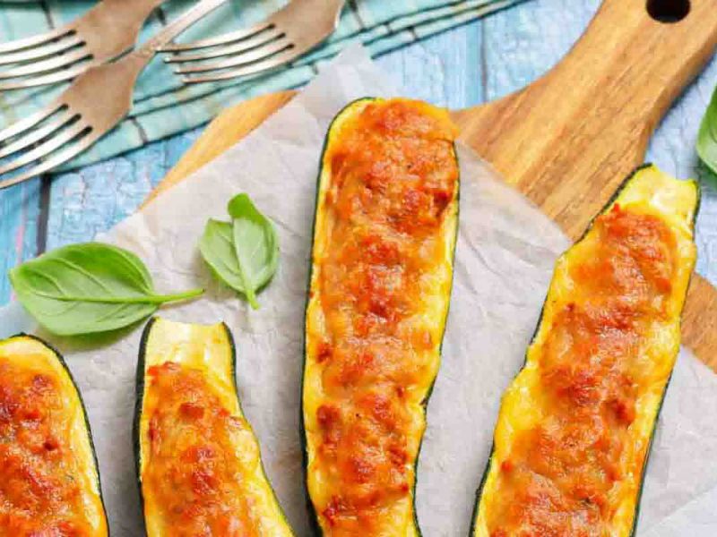 Four baked zucchini boats filled with melted cheese and a golden-brown topping are arranged on a parchment-lined wooden board, garnished with fresh basil leaves.