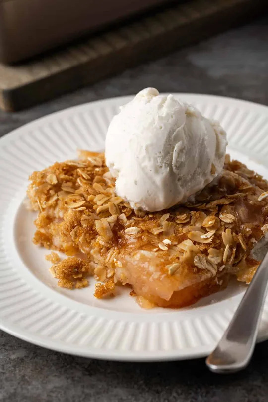 A slice of apple crumble topped with a scoop of vanilla ice cream on a white plate with a fork