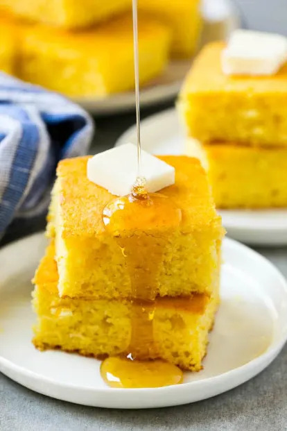 Stacked cornbread slices topped with butter and drizzled with honey on a white plate, with more cornbread in the background
