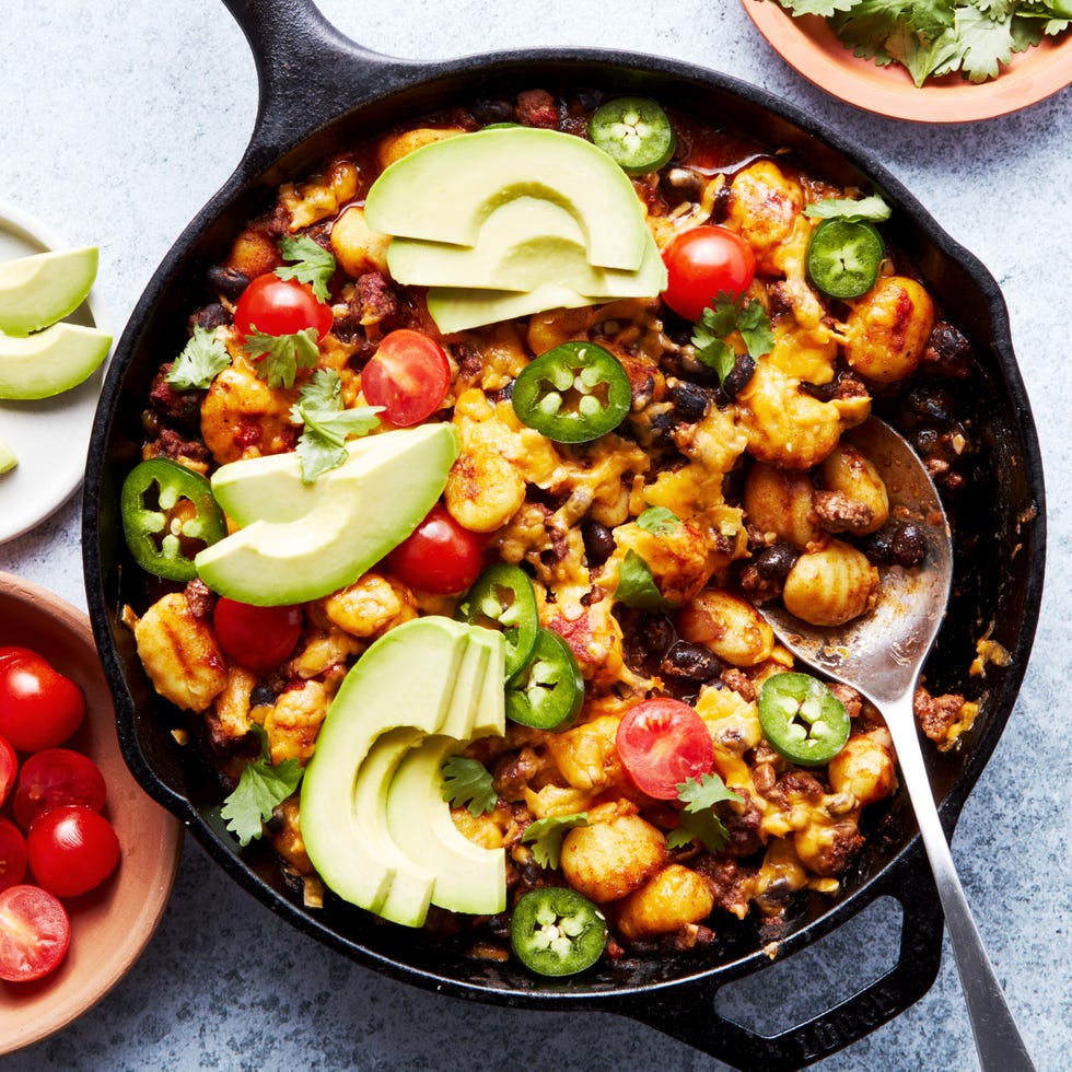 Taco Gnocchi Bake a pan with gnocchi covered in enchilada sauce, cheese, avocado, cilantro, and cherry tomatoes