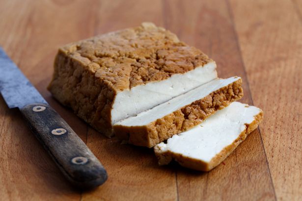 Block of smoked tofu, two tofu slices and rustic knife on wooden chopping board.