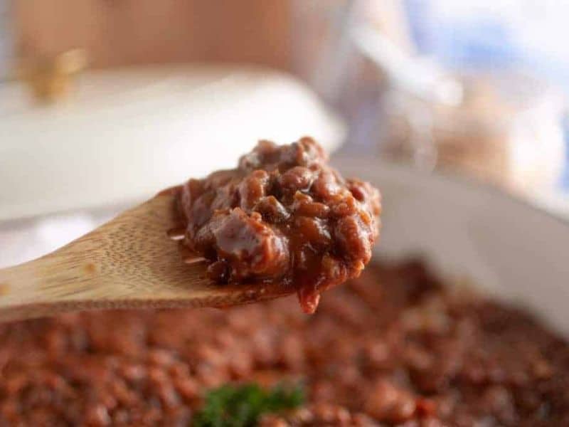 A wooden spoon holds a serving of chunky chili with beans and meat; a dish of chili is visible in the background.