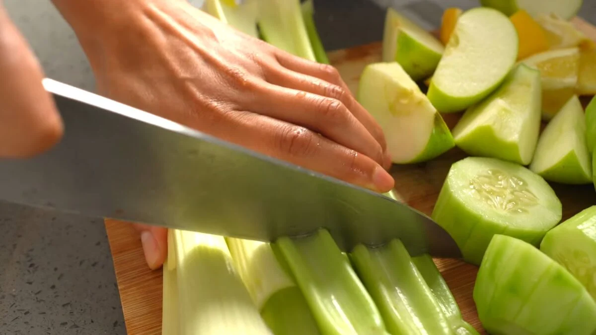 Reyes chops a bunch of celery on a chopping board next to sliced cucumber, apples, and lemon, to prep her favorite iron-rich green juice