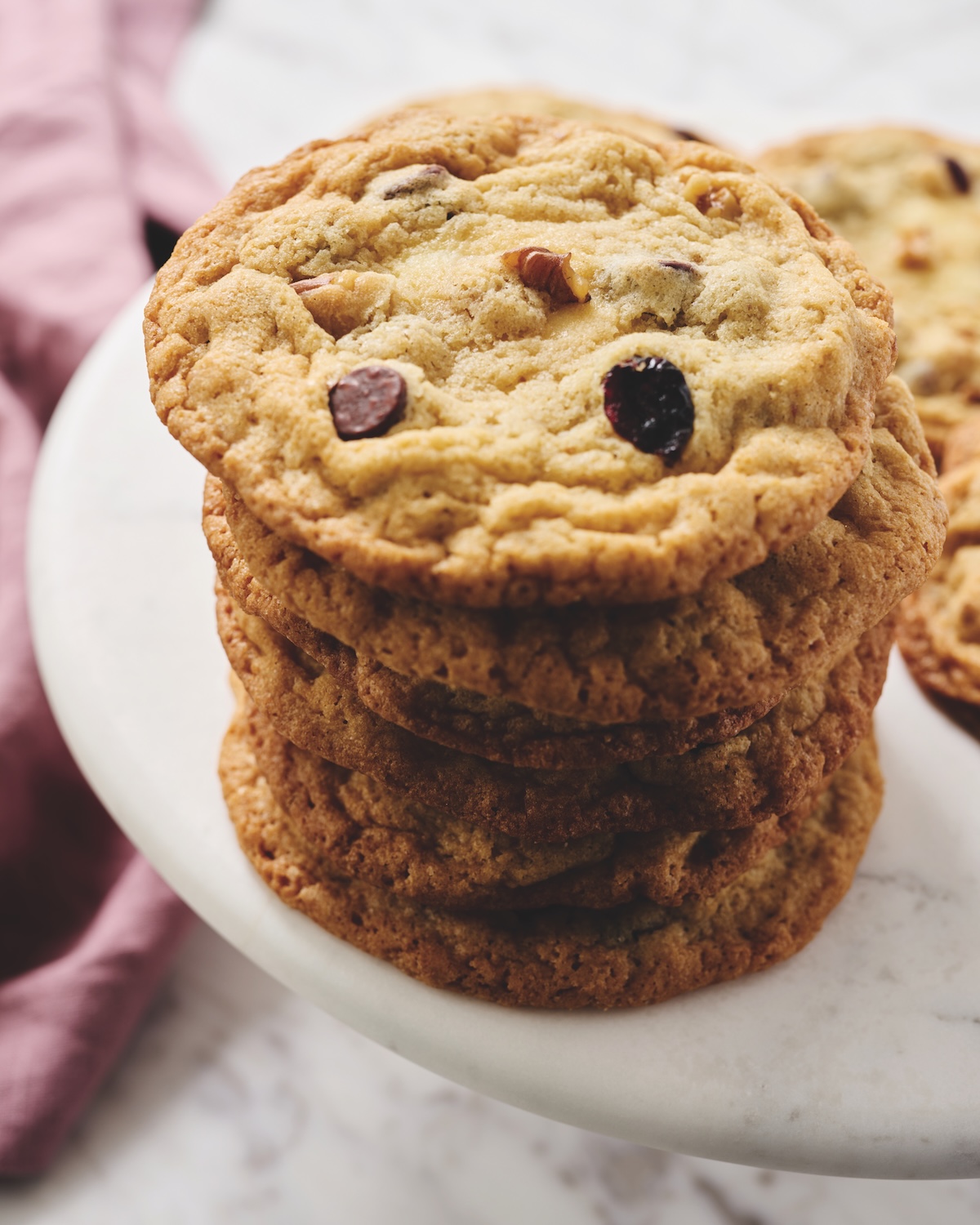A stack of chocolate chip cookies is arranged on a white marble plate, with a pink cloth visible in the background.