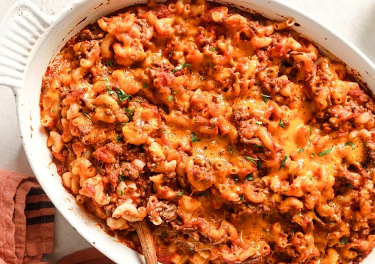 A close-up of a baked casserole dish filled with cheesy, saucy ground beef and macaroni, topped with melted cheddar cheese. A wooden spoon is partially visible in the dish.