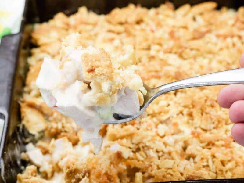A close-up of a fork holding a bite of creamy chicken casserole with a crumbly, golden topping above a baking dish of the same casserole.